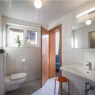 A modern bathroom with white tiles and a sink. There is a shower, a toilet, and a window that lets in natural light.