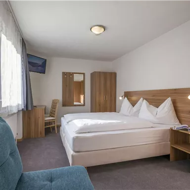 A modern hotel room with a double bed and a blue armchair. There is a window with daylight and a large mirror.