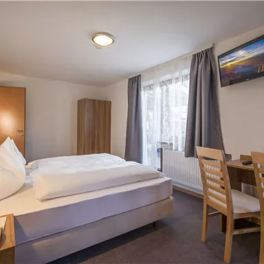 A cozy hotel room with a double bed, a desk, and a television. The windows provide natural light and a friendly atmosphere.