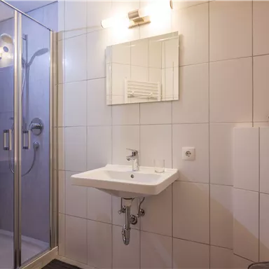 A modern bathroom with a shower cubicle and a sink. The walls are tiled with white tiles, and there is a mirror above the sink.