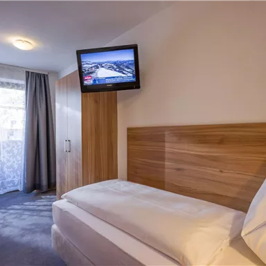 A simple bedroom with a single bed and a television on the wall. The room has a window with curtains and a bright, friendly atmosphere.