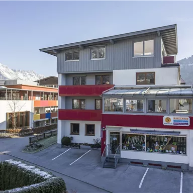 A modern building with a red and gray facade, surrounded by mountains. There is a parking lot and an entrance area with glass cladding.
