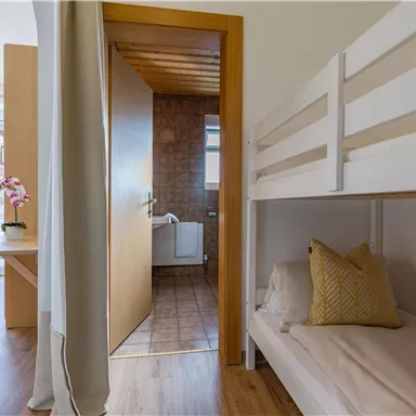 Ein modernes Zimmer mit einem Hochbett und einem gemütlichen Schlafbereich. Im Hintergrund ist ein Badezimmer und ein Tisch mit Stühlen zu sehen.