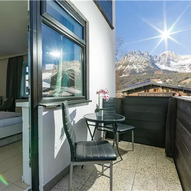 Ein Balkon mit einem Tisch und Stühlen, mit Blick auf majestätische Berge im Hintergrund. Die Sonne strahlt klar am Himmel und sorgt für eine einladende Atmosphäre.