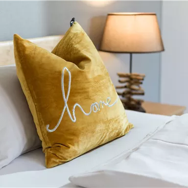 Ein gemütliches Schlafzimmer mit einem gelben Kissen, das das Wort "home" zeigt. Neben dem Bett steht eine Lampe, die eine warme Atmosphäre schafft.