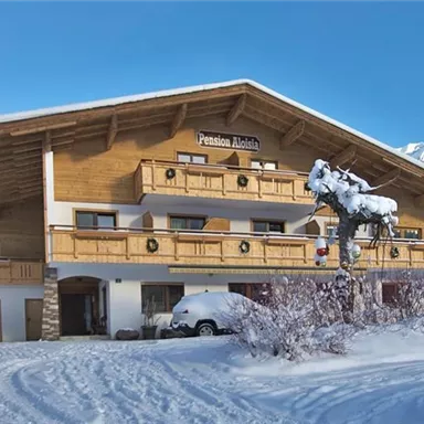 A cozy wooden house in the snow, surrounded by trees. The sky is clear and blue, creating a peaceful winter atmosphere.