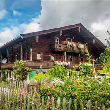 Ein traditionelles Holzhaus mit Balkonen, umgeben von bunten Blumen und grünen Pflanzen. Der Himmel ist bewölkt, und die Umgebung vermittelt eine ruhige ländliche Atmosphäre.