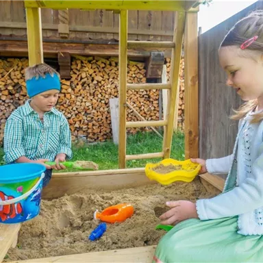 Zwei Kinder spielen in einer Sandkiste. Der Junge trägt ein grünes Hemd und eine blaue Mütze, während das Mädchen ein helles Kleid trägt und mit Sandspielzeug spielt.