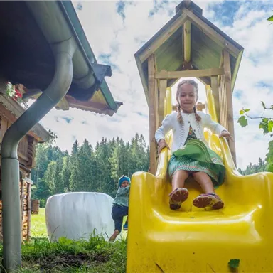 Ein Kind rutscht von einer gelben Rutsche in einem Garten. Im Hintergrund sind Bäume und ein Holzhaus zu sehen.