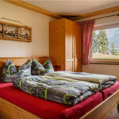 Ein gemütliches Schlafzimmer mit einem großen, roten Bett und zwei Kissen. Durch das Fenster ist der Blick auf die Natur sichtbar.