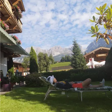 Eine ruhige Gartenlandschaft mit einem Liegestuhl und einer Person, die sich entspannt. Im Hintergrund sind Berge und ein blauer Himmel mit wenigen Wolken sichtbar.
