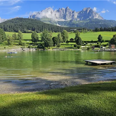 A quiet lake surrounded by green meadows and forests. Majestic mountains rise in the background under a blue sky.