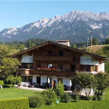 Ein malerisches Chalet umgeben von grünem Rasen und Bäumen. Im Hintergrund sind beeindruckende Berge sichtbar.