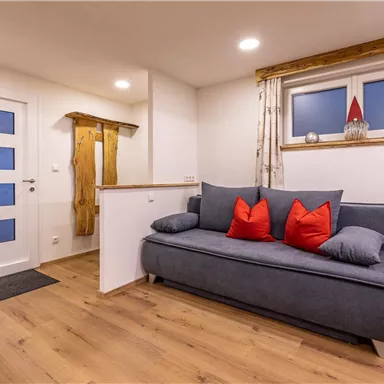 A modern living room with a gray sofa and red cushions. The walls are bright and inviting, and there are large windows for daylight.