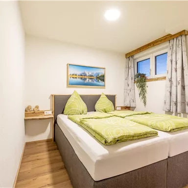 A bright bedroom with a double bed and green cushions. A landscape painting hangs on the wall and a small window lets light in.