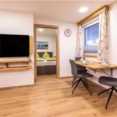 A cozy living space with a TV, a dining table, and two chairs. The room has light-colored walls and a wooden floor.