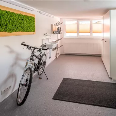 Ein minimalistischer Raum mit einem Fahrrad, hellen Wänden und einem grünen Moosbild an der Wand. Es gibt einen Schrank und Fenster, die natürlichen Licht einlassen.