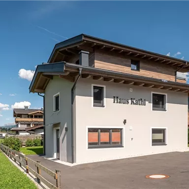 Ein modernes Haus mit dem Namen "Haus Kathl" in einer grünen Umgebung. Die Fassade ist hell und freundlich, umgeben von Wiesen und Bergen.