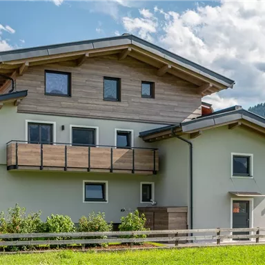 Ein modernes Haus mit einer Holzverkleidung und großen Fenstern. Umgeben von Grünflächen und unter einem klaren Himmel.