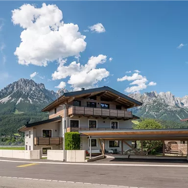 Ein modernes Haus mit einem Holzcarport vor einer malerischen Berglandschaft. Der Himmel ist blau mit einigen Wolken.