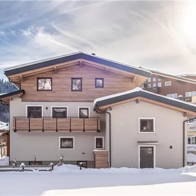 Ein modernes Haus im Schnee mit einer Holzveranda. Im Hintergrund sind Berge und weitere Gebäude zu sehen.