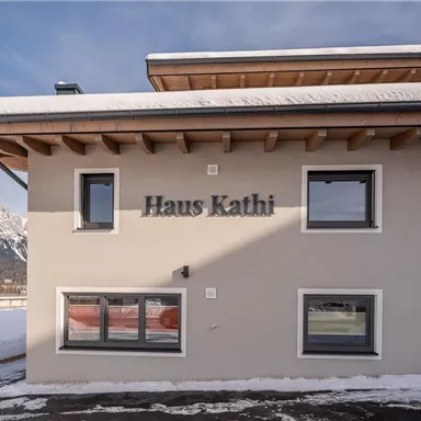 Ein modernes Gebäude mit der Beschriftung "Haus Kathi". Es liegt in einer schneebedeckten Landschaft mit Bergen im Hintergrund.