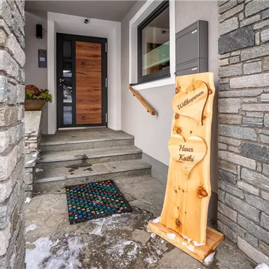 Eingang zu einem modernen Haus mit einer Holztür und einem begrüßten Fußabtreter. Neben der Tür steht ein Willkommensschild mit der Aufschrift "Haus Kathi".