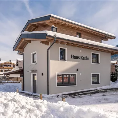 Ein modernes Haus im Schnee mit dem Namen "Haus Kathi" an der Fassade. Im Hintergrund sind Berge und weitere Gebäude zu sehen.
