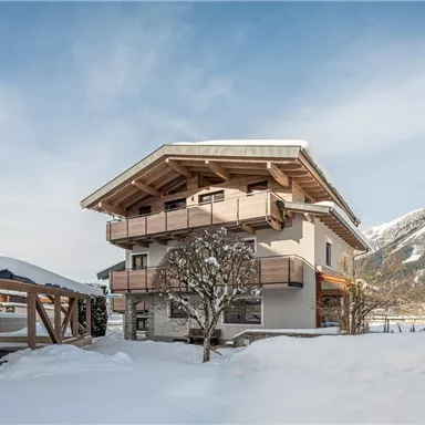 Ein modernes Haus im Schnee mit Holzverkleidung und Balkonen. Im Hintergrund sind Berge unter einem klaren Himmel zu sehen.