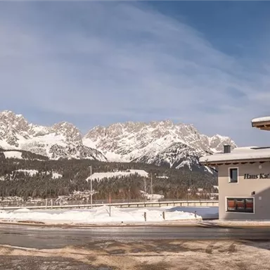 Eine malerische Winterlandschaft mit schneebedeckten Bergen im Hintergrund. Im Vordergrund steht ein modernes Gebäude an einer gut ausgebauten Straße.