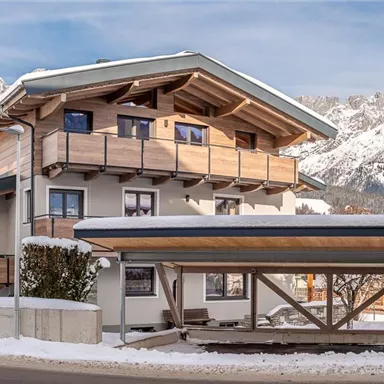 Ein modernes Haus im alpinen Stil, umgeben von Schnee und Bergen. Die Fassade aus Holz und der Balkon verleihen dem Gebäude einen einladenden Charakter.