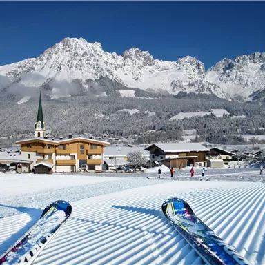Eine schneebedeckte Landschaft mit Bergen im Hintergrund. Im Vordergrund befinden sich Skier auf der präparierten Piste und ein schöner Ort mit traditionellen Gebäuden.