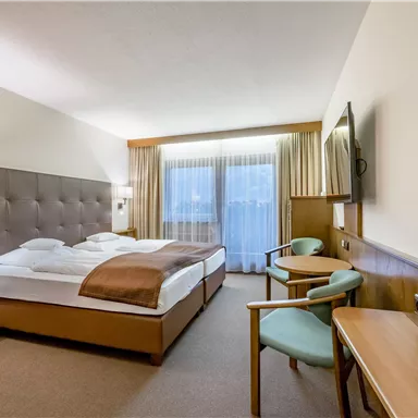 Ein modernes Hotelzimmer mit einem großen Bett, einem Fernseher und einem kleinen Tisch. Durch die Fenster scheint Tageslicht, und es gibt zwei Stühle neben dem Tisch.