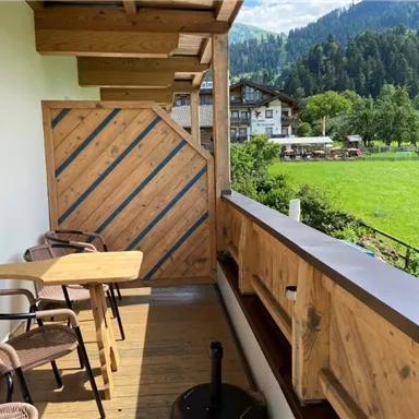 Ein Holz-Balkon mit zwei Stühlen und einem Tisch. Im Hintergrund sieht man eine grüne Wiese und Berge.