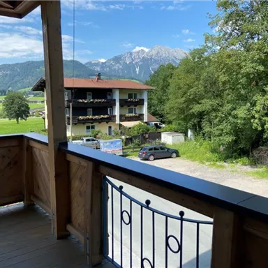 Blick von einem Balkon auf eine grüne Landschaft mit Bergen im Hintergrund. Im Vordergrund steht ein Holzgeländer und ein Gebäude ist zu sehen.