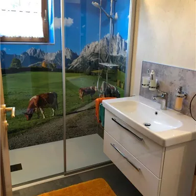 Ein modernes Badezimmer mit einer Dusche und einem Waschtisch. Die Duschwand zeigt ein malerisches Bergpanorama mit Kühen auf einer Wiese.