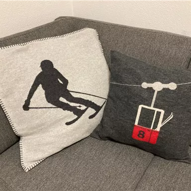 Zwei dekorative Kissen auf einem grauen Sofa. Eines zeigt einen Skifahrer und das andere eine Skilift-Gondel mit der Nummer 8.