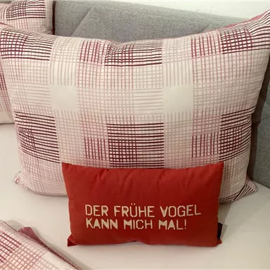 Ein großes Kissen mit einem geometrischen Muster und ein rotes Kissen mit dem Spruch „DER FRÜHE VOGEL KANN MICH MAL!“ auf einem Bett. Die Kissen verleihen dem Raum eine gemütliche Atmosphäre.