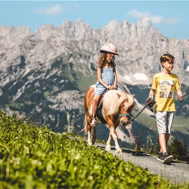 A girl is riding a pony while a boy is leading it by a rope. Impressive mountains can be seen in the background.