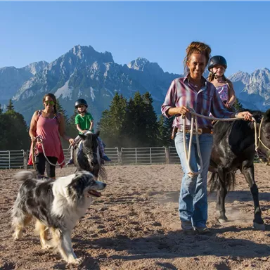 Eine Frau führt ein Pony, während Kinder mit einem anderen Pony und einem Hund spielen. Im Hintergrund sind Berge und Bäume zu sehen.
