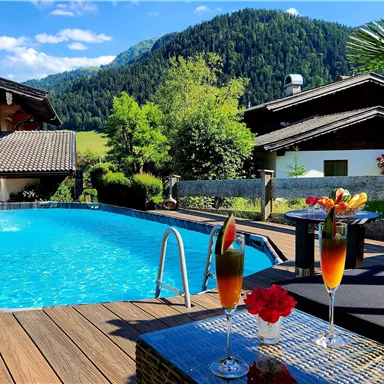 Ein schöner Außenpool umgeben von einer Holzterrasse und üppigem Grün. Im Vordergrund stehen zwei Cocktails mit frischen Früchten.