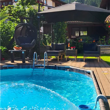 Ein klarer Pool in einem schönen Garten, umgeben von Pflanzen und einer attraktiven Sitzgelegenheit. Im Hintergrund sind Berge und blauer Himmel zu sehen.