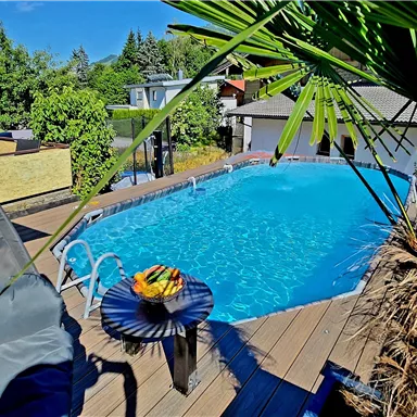 Ein einladender Pool umgeben von Pflanzen und einer Holzterrasse. Auf dem Tisch liegt eine Schale mit frischem Obst.