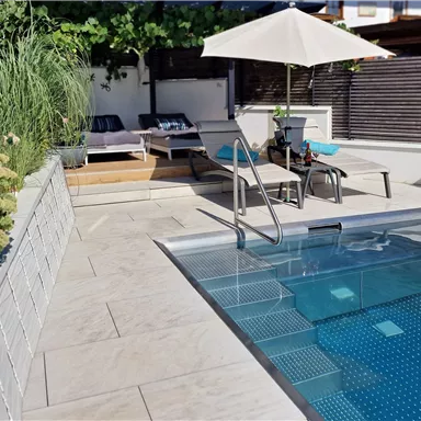 A modern garden with a small pool and lounge chairs. Next to the pool is a sun umbrella, and there are green plants in bowls.