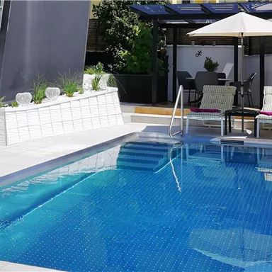 A beautiful pool with clear water in a modern garden. In the background, there are lounge chairs and a sun umbrella.