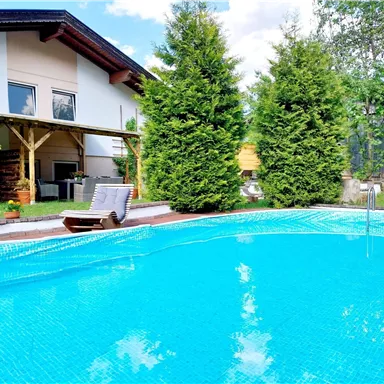 A clear pool surrounded by green trees and a cozy garden. In the background, there is a modern house with a covered terrace.