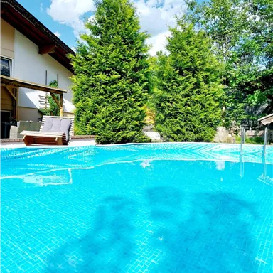 A clear pool surrounded by green trees and a modern house. The sun is shining and the sky is blue with some clouds.