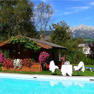Ein gemütliches Holzhaus mit bunten Blumen steht neben einem Swimmingpool. Im Hintergrund sind die majestätischen Berge zu sehen.