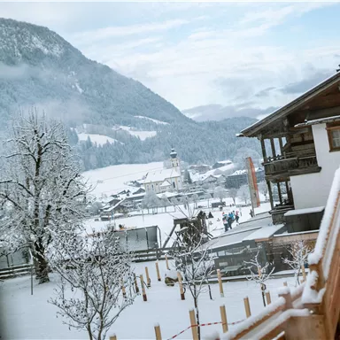 Eine verschneite Landschaft mit Bergen im Hintergrund und traditionellen Holzgebäuden. Die Szene strahlt eine ruhige Winteratmosphäre aus.