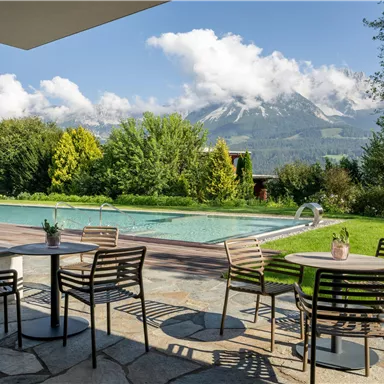 Ein schöner Außenbereich mit Stühlen und Tischen. Im Hintergrund befindet sich ein Swimmingpool und majestätische Berge unter einem blauen Himmel.